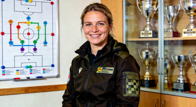 莱娜·沃格特 Lena Vogt 自信微笑，身穿教练夹克，背景是战术白板与奖杯柜